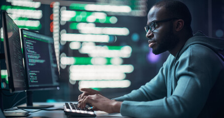 Black Male Programmer Working in Monitoring Room, Surrounded by Big Screens Displaying Lines of...
