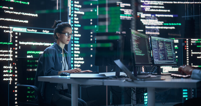 Portrait of a Woman Working on a Computer, Typing Lines of Code that Appear on Big Screens Surrounding her in a Monitoring Room. Female Programmer Creating Innovative Software Using AI Data and System