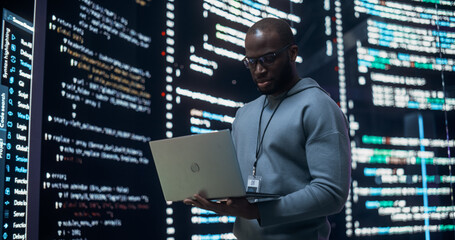 Portrait of Young Black Man Working on Laptop, Looking at Big Digital Screen Displaying Back-end Code Lines. Professional Programmer Developing a Big Data Interface Software Project Concept.