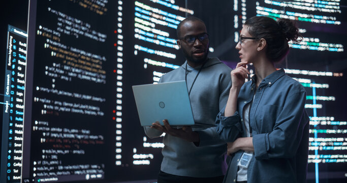 Portrait of Two Diverse Developers Using Laptop Computer, Discussing Lines of Code that Appear on Big Screens Surrounding Them. Male and Female Programmers Creating a Software Together and Fixing Bugs