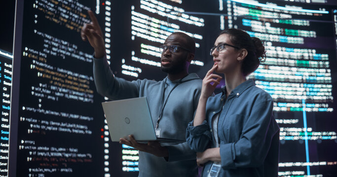Portrait of Two Diverse Developers Using Laptop Computer, Discussing Lines of Code that Appear on Big Screens Surrounding Them. Male and Female Programmers Creating Software Together and Fixing Bugs