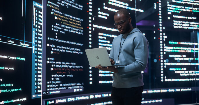 Portrait of Young Black Man Working on Laptop Computer, Looking at Big Digital Screen Displaying Back-end Code Lines. Professional Programmer Developing a Big Data Interface Software Project