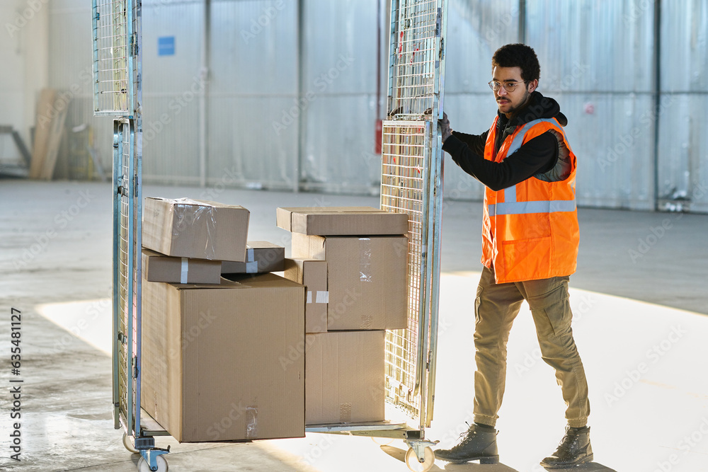 Poster young worker of warehouse in workwear pushing cart with stack of packed cardboard boxes containing s