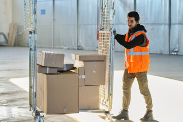 Young worker of warehouse in workwear pushing cart with stack of packed cardboard boxes containing spare parts for industrial equipment © pressmaster