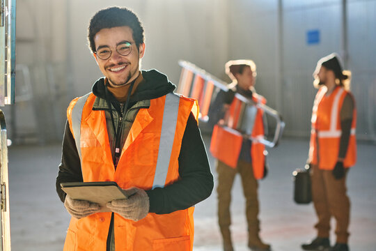 Young successful male technician looking at camera and using tablet while looking through online manual against two colleagues in warehouse
