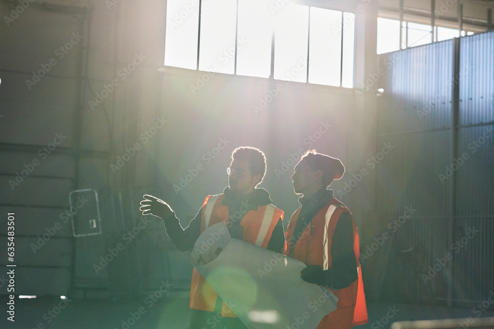 Poster Sunlit outlines of two young intercultural engineers consulting about sketch points while one of them holding blueprint with building plan