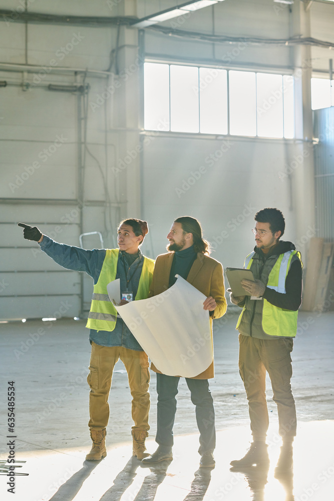 Poster young confident male engineer pointing at wall of warehouse during discussion of working points and 