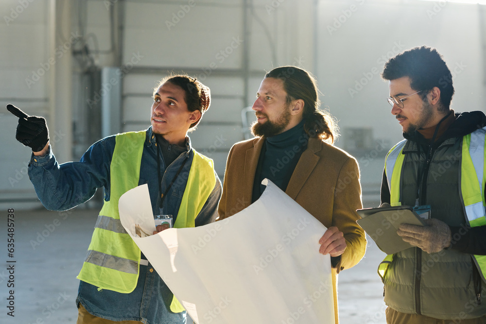 Poster young engineer and contractor with blueprint listening to warehouse worker pointing at something dur