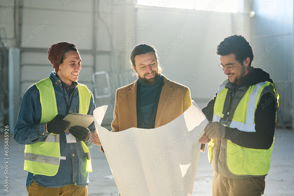 Poster happy young contractor and two employees in safety jackets looking at sketch or building plan on blu