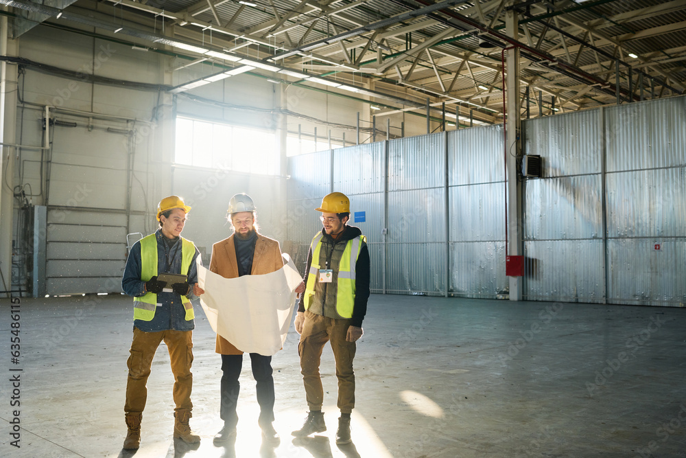 Poster Three engineers in protective helmets discussing blueprint with building plan while standing in warehouse against sunlight coming through window