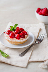 Cake with cream and raspberries decorated with a mint leaf, napkin, cutlery, bowl with raspberries, on a light background.