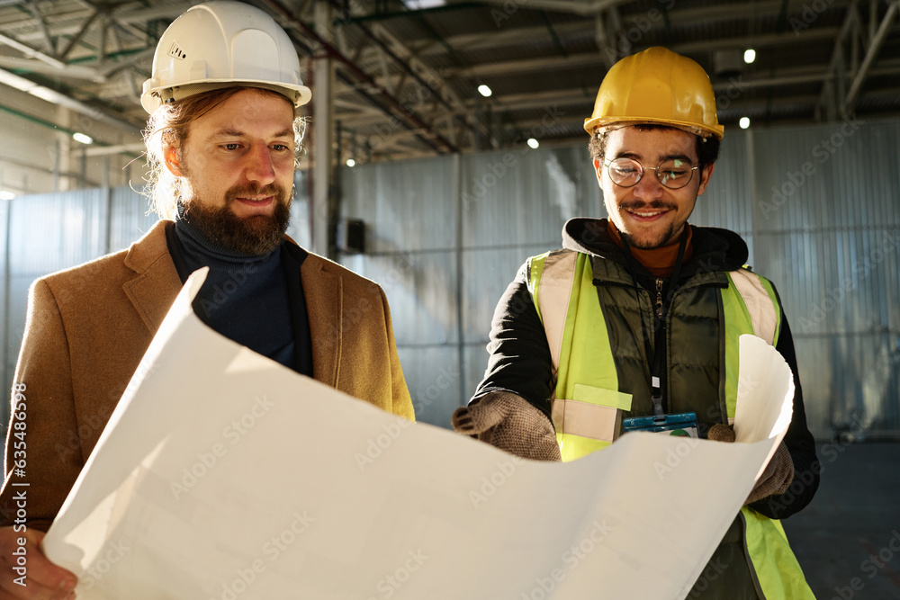 Poster Young smiling engineer explaining details of building plan to contractor or foreman in hardhat while pointing at blueprint at working meeting