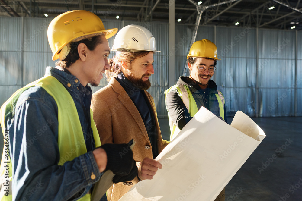 Aufkleber Young confident manager in protective helmet and reflective vest explaining sketch on blueprint while standing next to contractor