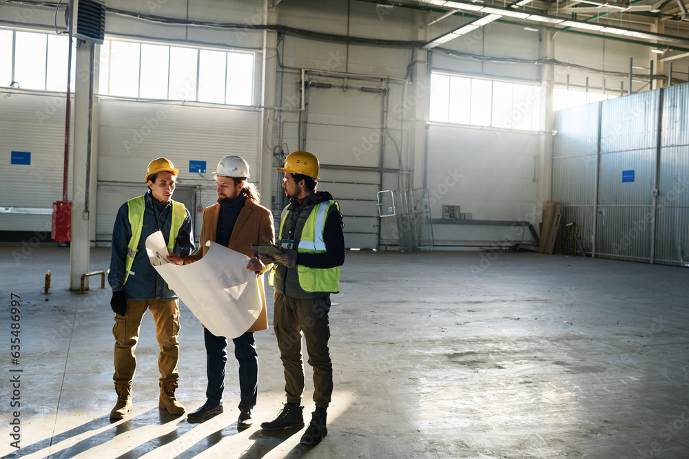 Poster long shot of two young multiethnic engineers in protective workwear and contractor with blueprint lo
