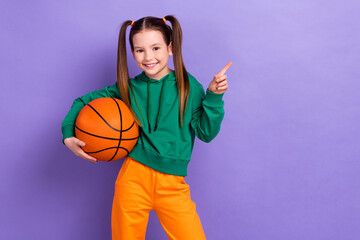 Photo of positive sweet small girl wear green sweatshirt playing basketball pointing empty space isolated violet color background