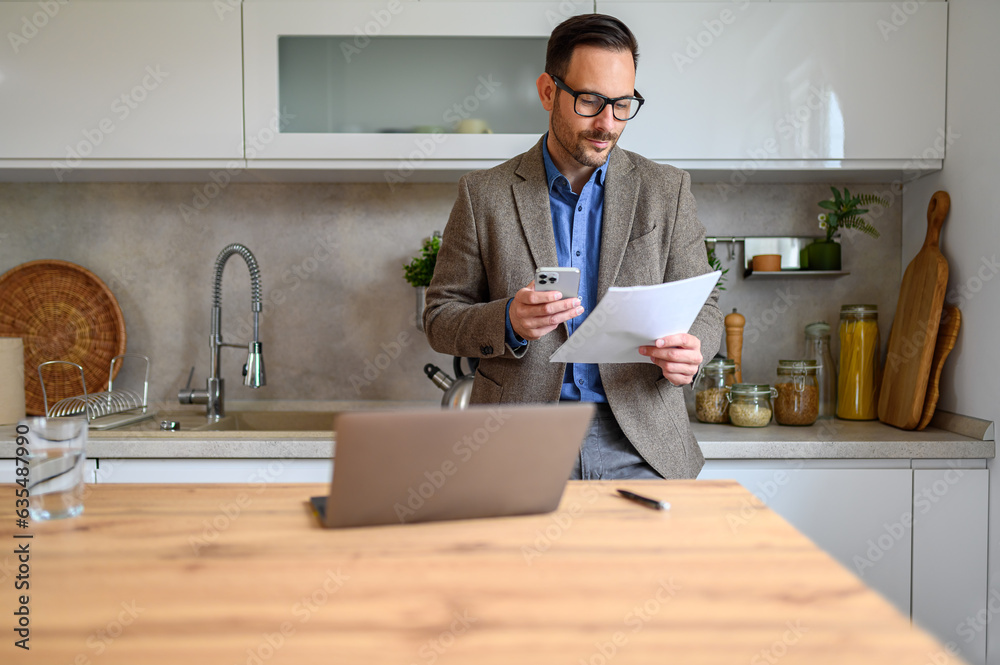 Wall mural elegant businessman in suit holding report and messaging on cellphone while standing in home office