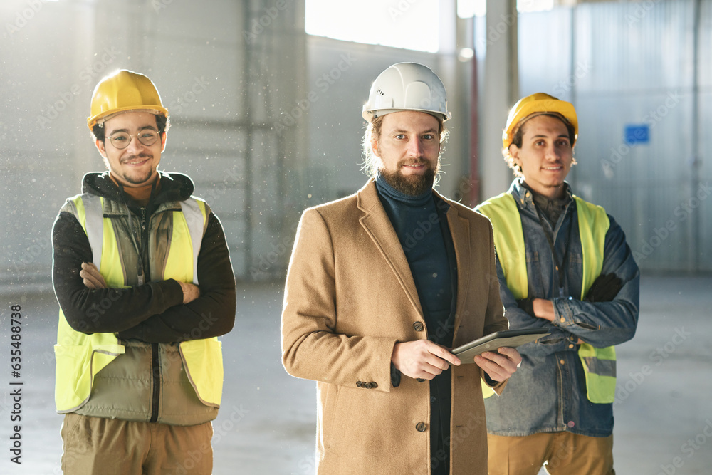 Poster happy young foreman in beige coat and two multirthnic engineers in protective helmets and safety ref