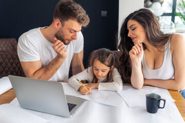 Young parents teach their daughter to write, helping her with her homework, untidy desk and laptop. Concept happy healthy family