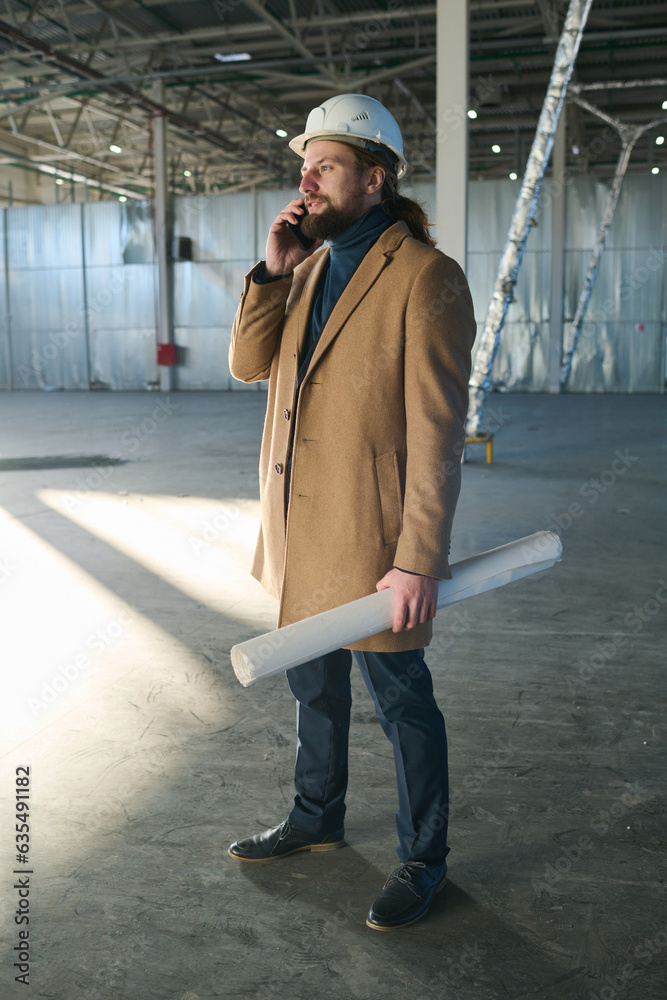 Poster side view of young bearded engineer in casualwear and protective helmet standing in front of camera 