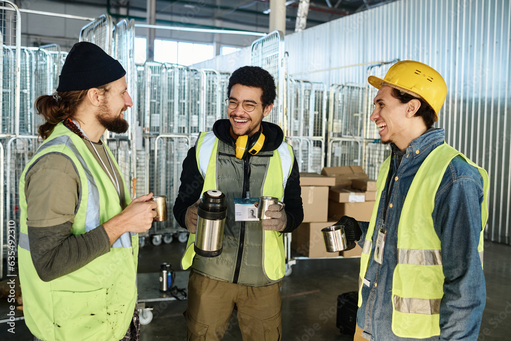 Poster Three young cheerful engineers in workwear looking at each other while discussing something funny and having hot tea at break in warehouse