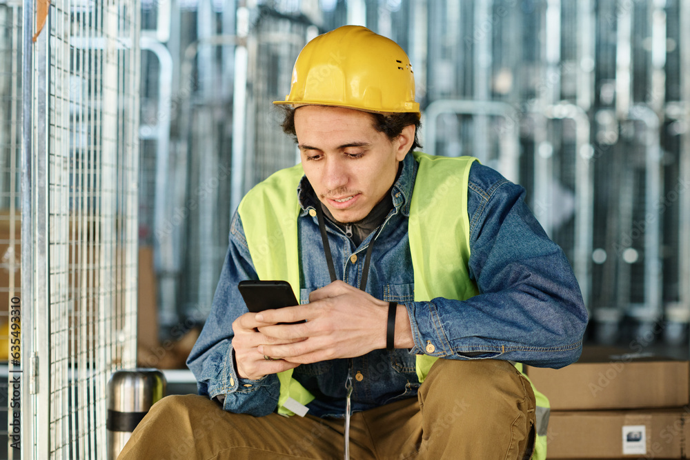Poster young male engineer in workwear and safety helmet texting in smartphone while sitting on large metal