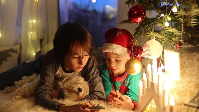 Toddler child, cute blond boy, sitting on the window in pajama, looking out for Satna Claus,, eating cookies,  Christmas lights around him