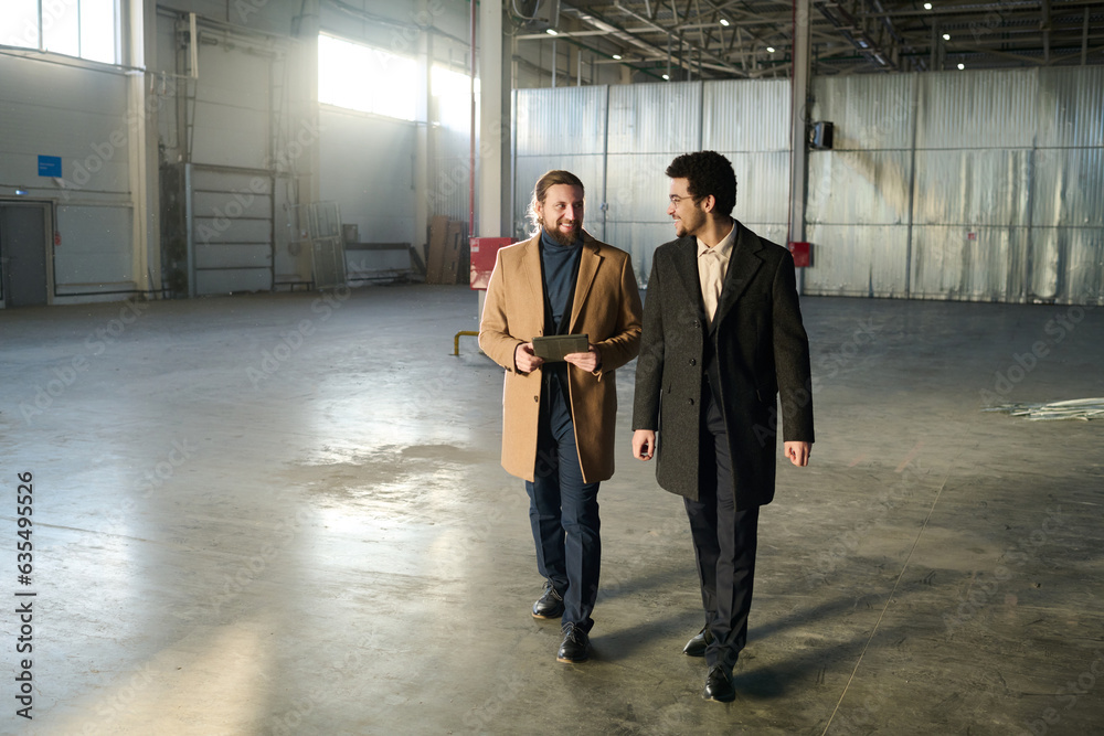 Poster two young successful entrepreneurs in elegant coats moving along spacious empty warehouse and discus