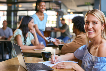 Portrait Of Woman Working On Laptop Sitting At Table In Busy Coffee Shop