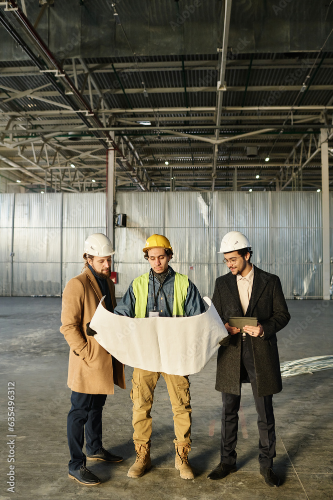 Leinwandbilder Front view of team of young architects or engineers discussing building plan or sketch on unrolled blueprint held by worker in safety vest
