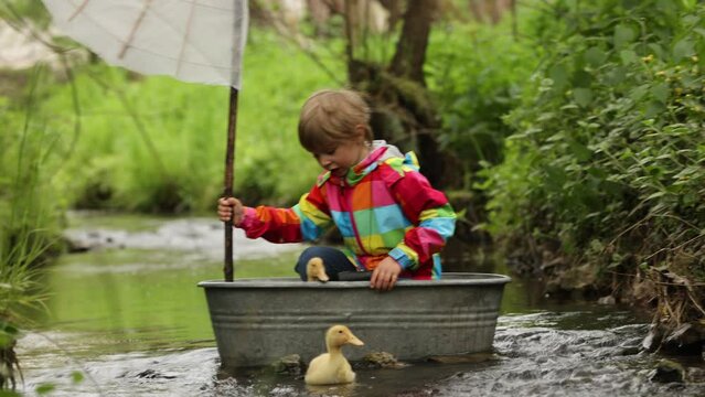 Cute child, boy in colorful jacket, playing with boat and ducks on a little river, sailing and boating. Kid having fun, childhood happiness concept