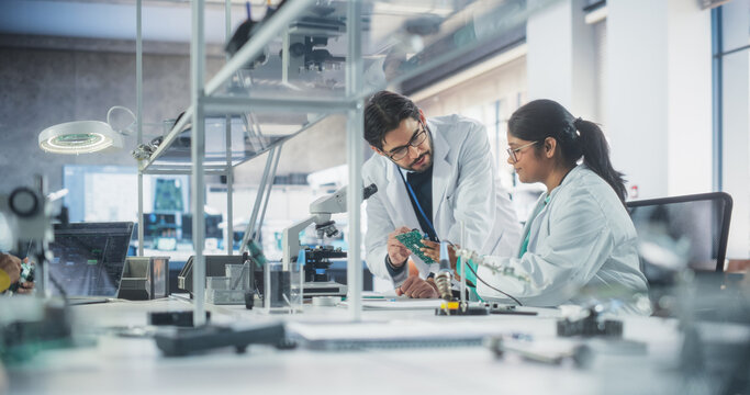 Two Diverse Multiethnic Colleagues Chatting in Laboratory, Working on a Circuit Board for a Computer. Young Indian Scholar Talking with a Scientist About Inserting Different Microchips and Conductors - Powered by Adobe