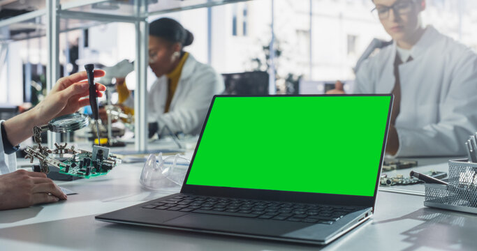 Laptop Computer with Green Screen Mock Up Display Standing on a Table in a Science Class Laboratory. Diverse Students Practicing Soldering Electronic Components for Their Tech Projects