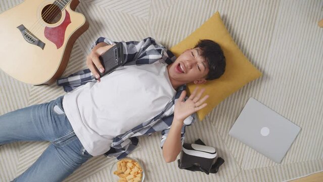 Top View Of Asian Teen Boy Having Video Call On Smartphone While Lying On Carpet On The Floor At Home. Waving Hands, Smiling, And Speaking
