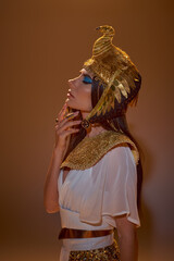 Woman in Egyptian attire and headdress touching chin while posing on brown background