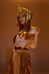 Woman in egyptian attire pointing with finger at camera and standing on brown background with light