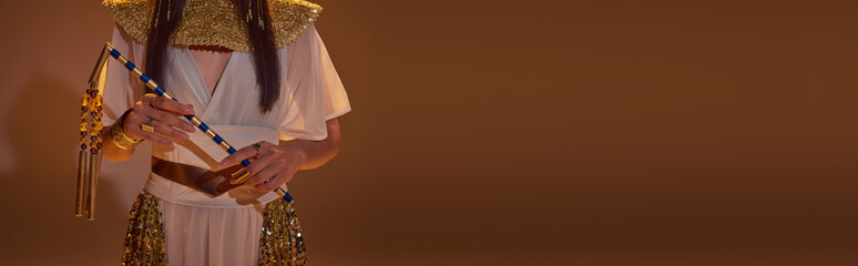 Cropped view of woman in elegant egyptian look holding flail on brown background, banner