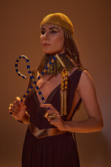 Elegant woman in egyptian golden headdress and look holding crook and flail on brown background