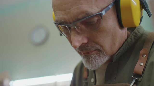 Tilt up of mature woodworking master in apron, eyeglasses and protective headphones holding electric sander and polishing surface of wooden workpiece while standing by workbench