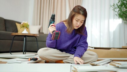 Young asian women holding a drill for assembling new furniture by herself at home. Woman sitting on floor in living room holding a drill for assembling furniture. Assembling furniture concept