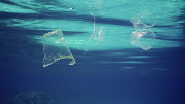 Close-up of plastic debris drifts under surface in blue water, Slow motion. Disposable plastic cups and pacs littering oceans of planet. Ecological problems, defending environment from pollution. 