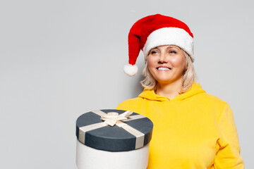 Studio portrait of smiling woman in yellow sweater wearing Santa hat, holding gift in hand, on white background.