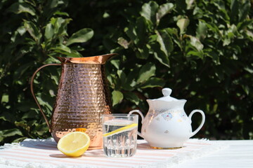 Copper pitcher and water glass with lemon on the table, outdoor. Summer still life in vintage style.