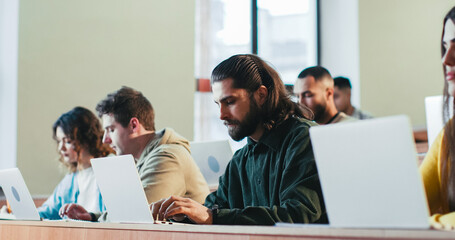 Caucasian guy student asing something to professor. Students sitting at desks with laptops and studying. Indoor. University concept. Young adults learning at seminar in college. Knowledge gaining.