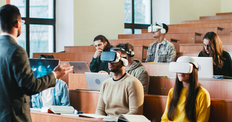 Rear of professor having speech in front of mixed-races students in VR glasses and laptops. Multiethnic males and females studying in technologic high school and listening to teacher at seminar.