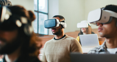 Close up of mixed-races males and female students in VR glasses listening to lection at college and rising hands to answer. Hi-tech study. Multiethnic young people having virtual reality education.