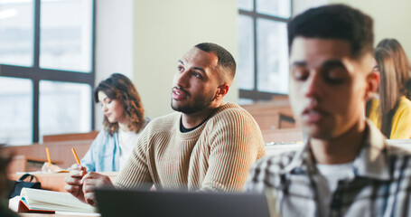 Young African American male student sitting among other students at lection and asking question to professor. Indoor. Man communicating with teacher at seminar in college. Studying concept.