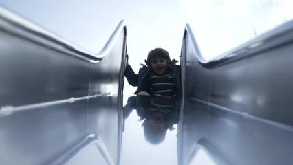 Joyful child sliding down slider at playground park during fall season, kid wearing jacket with smile on face going down toward camera