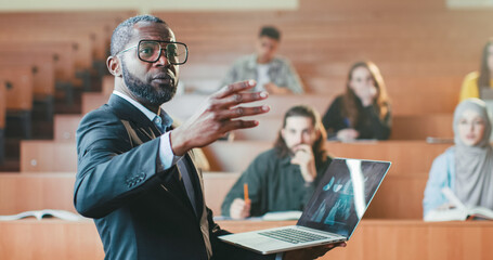 African American male professor standing in front of students in college with laptop and talking to...