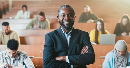 Portrait shot of handsome happy African American male professor crossing hands at University, looking at camera and laughing. Students in class on background. Man teacher of high school. Study concept