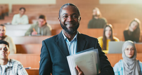 Portrait of happy African American male professor smiling in front of camera and holding folder with papers. Students on background. Man teacher of high school. Camera zooming out. Close up.